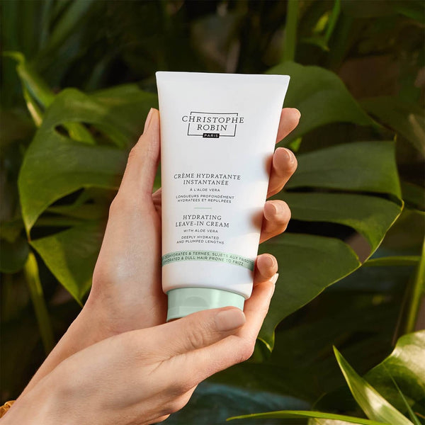 Christophe Robin Hydrating Leave-In Cream With Aloe Vera held in the palm of a hand in front of a palm tree
