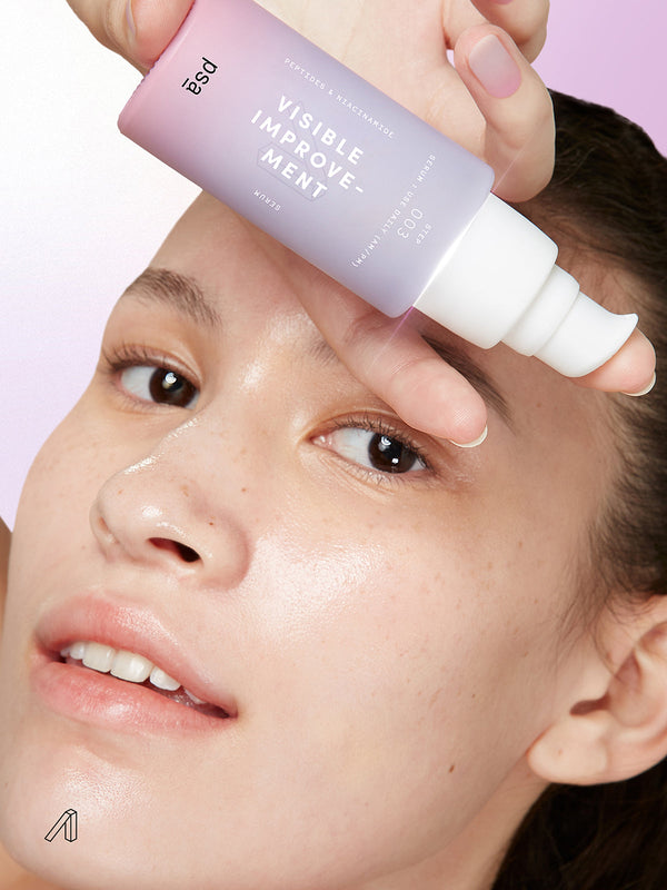 a closeup of a person holding a bottle of PSA VISIBLE IMPROVEMENT Peptides & Niacinamide Serum close to their face